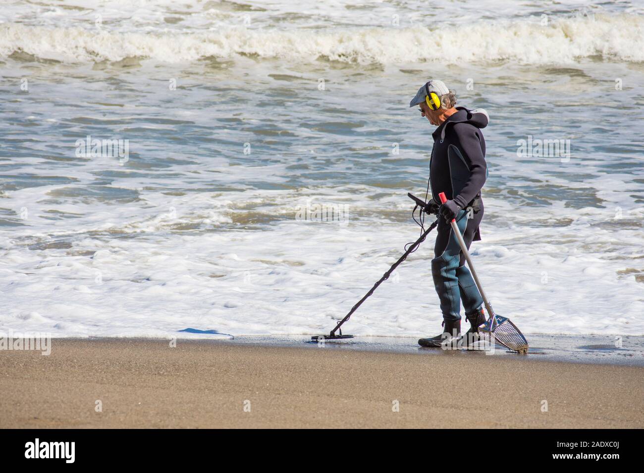 Hombre en wet suit buscando en la playa con el detector de metales, España. Foto de stock