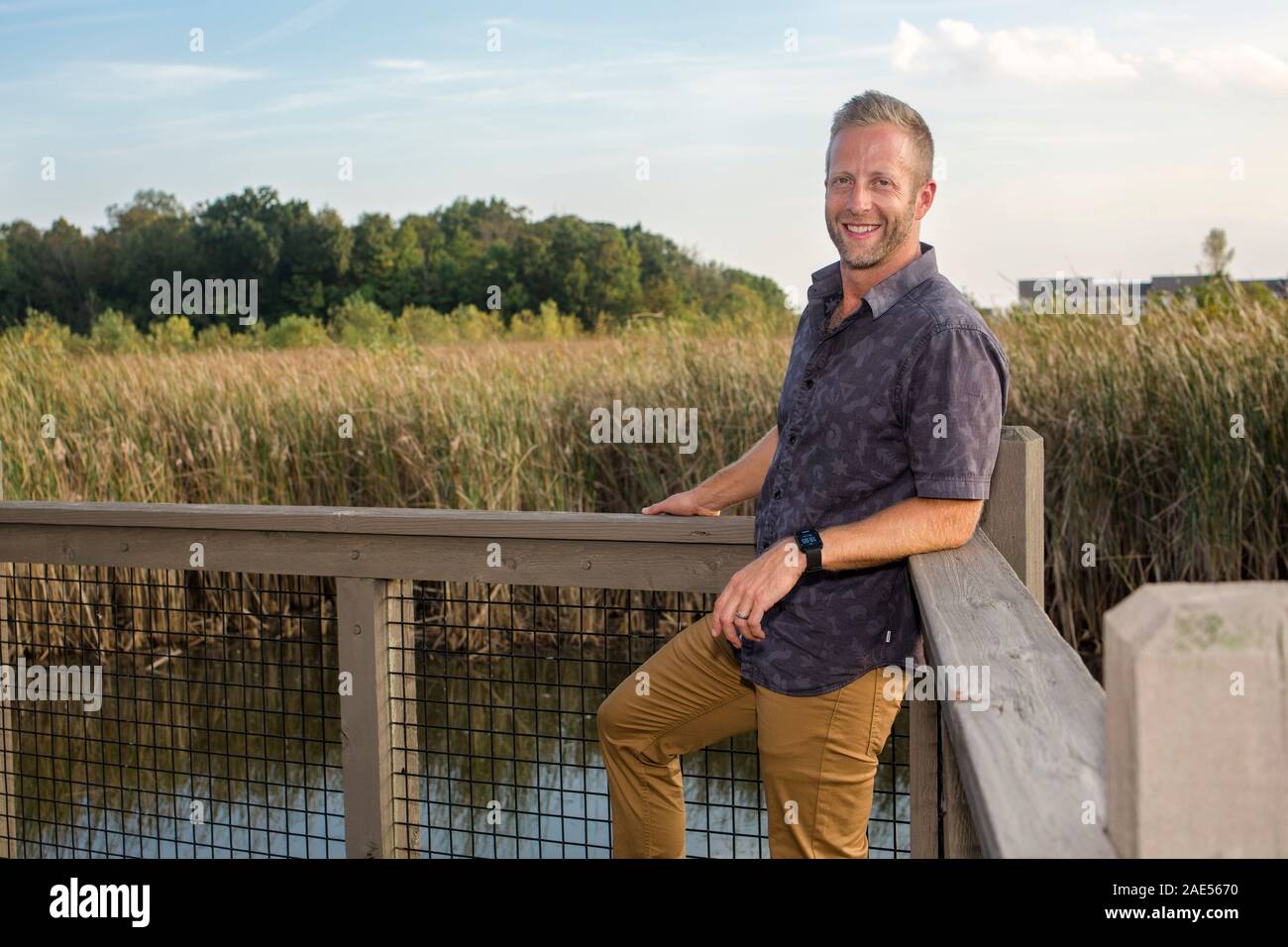Un guapo hombre sonriente se inclina contra una barandilla en un parque de humedales Foto de stock