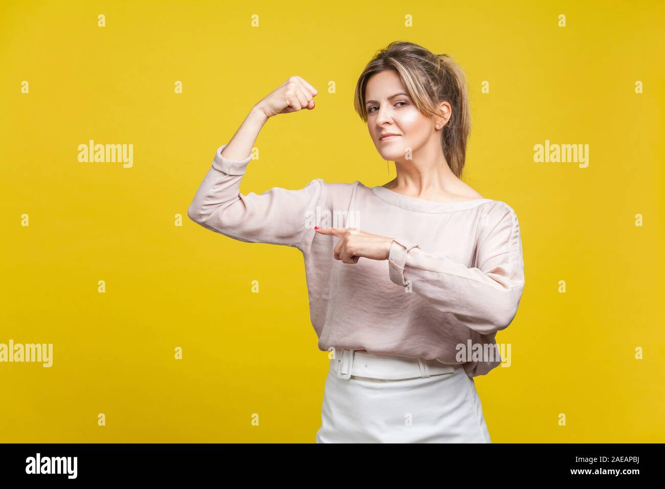 Soy fuerte. Retrato de mujer poderosa en blusa casual de pie, señalando a su mano como mostrando bíceps, buscando seguros e independientes del interior. Foto de stock
