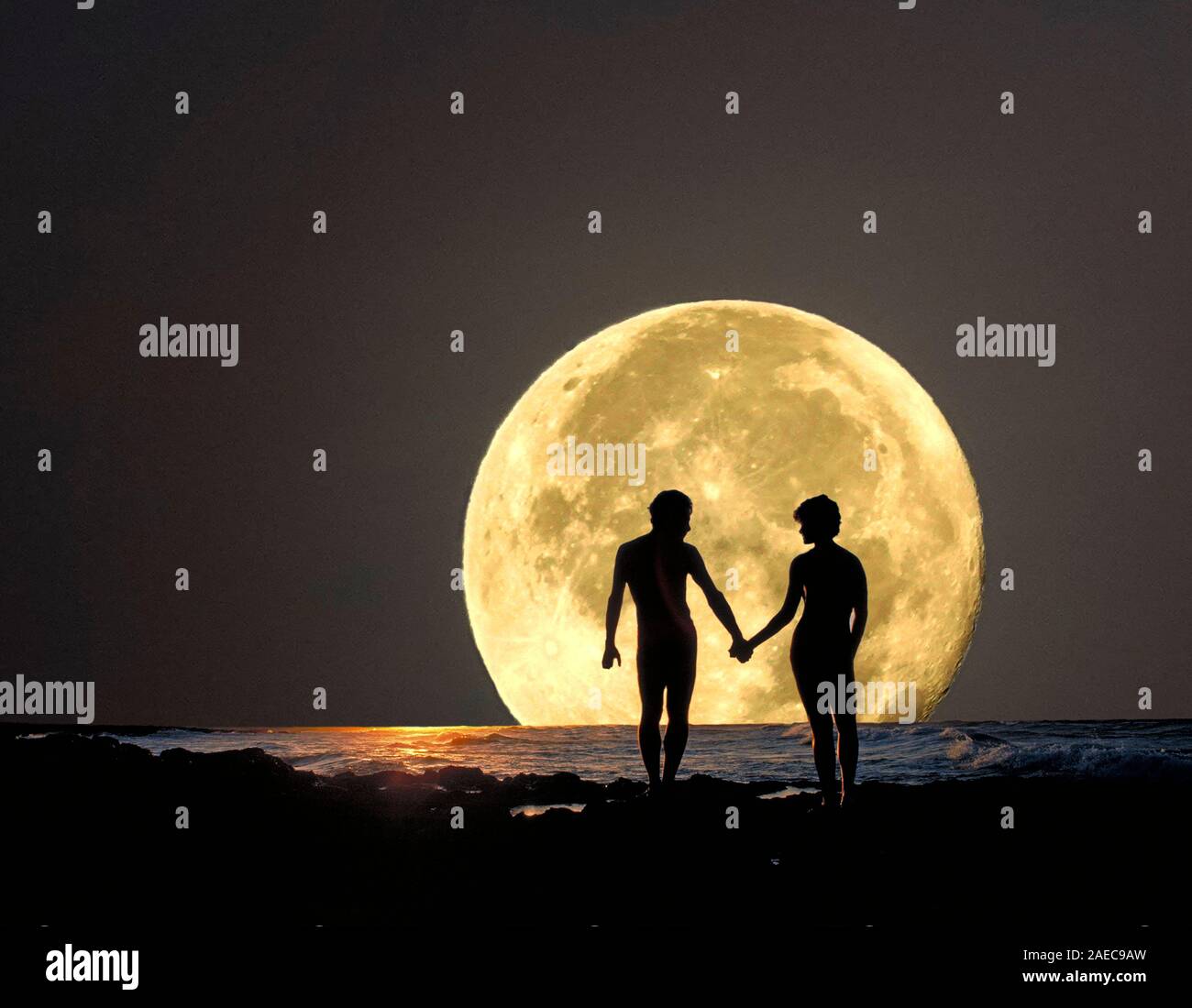 Una joven pareja que sostiene las manos sobre una zona rocosa en la costa del Océano Pacífico, mientras que la luna llena se eleva sobre el horizonte. Foto de stock