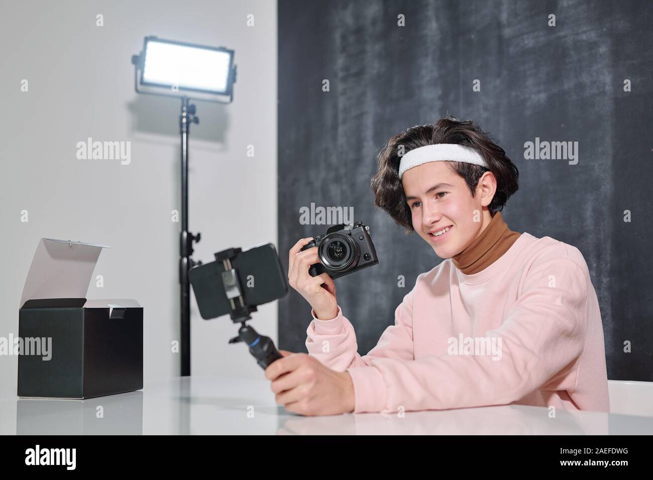 Sonriente joven varón vlogger con photocamera sentado al escritorio y disparar a sí mismo Foto de stock