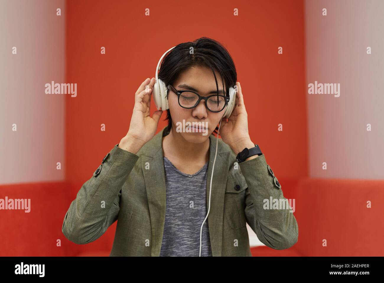 Joven asiática en los anteojos que lleva puestos los auriculares y escuchar música con fondo naranja Foto de stock