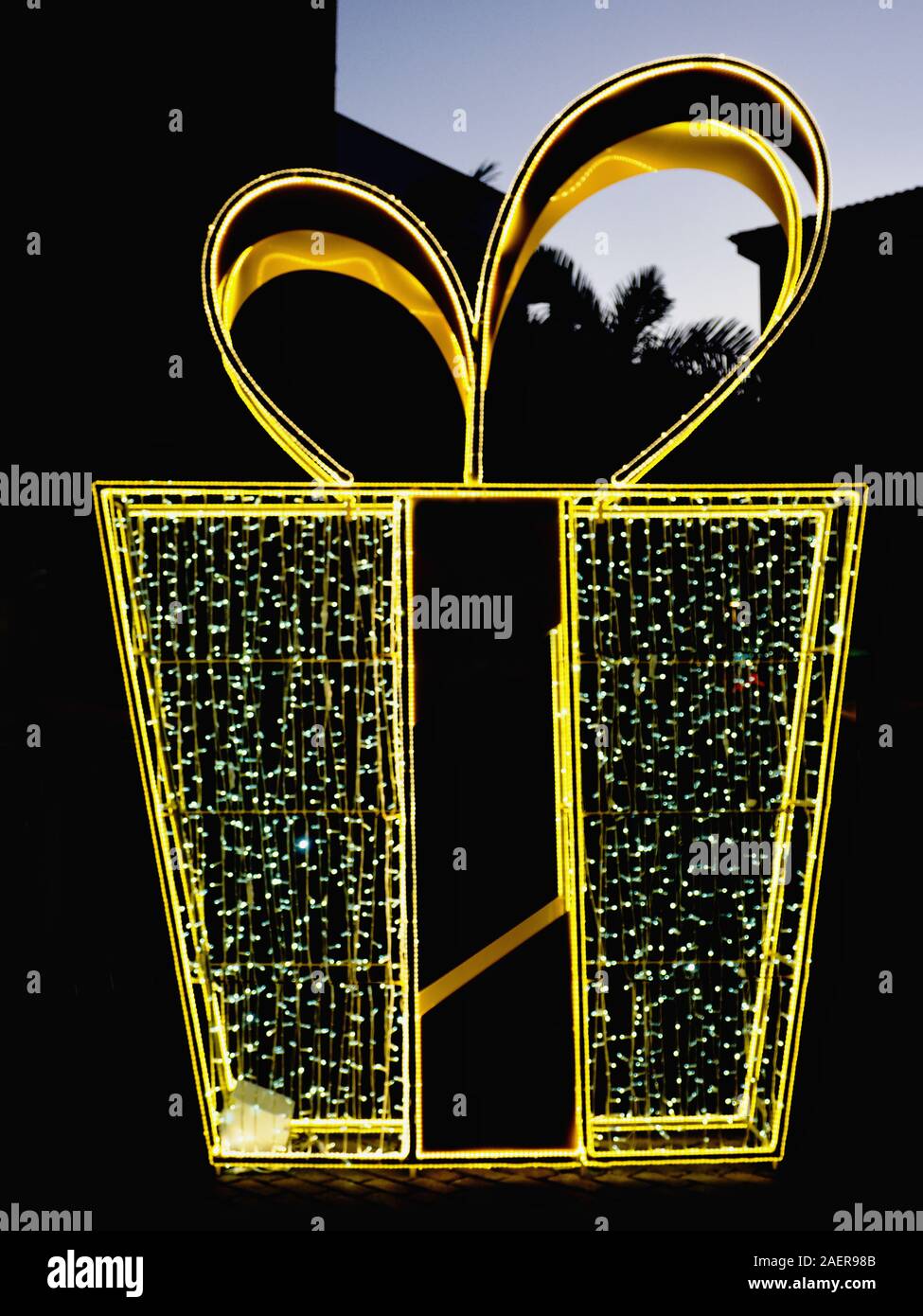 Una gran decoración de Navidad en una ciudad en la oscuridad, en la forma de una enorme caja de regalo en oro y las luces de hadas y una cinta ancha en la parte superior. Close-up i Foto de stock