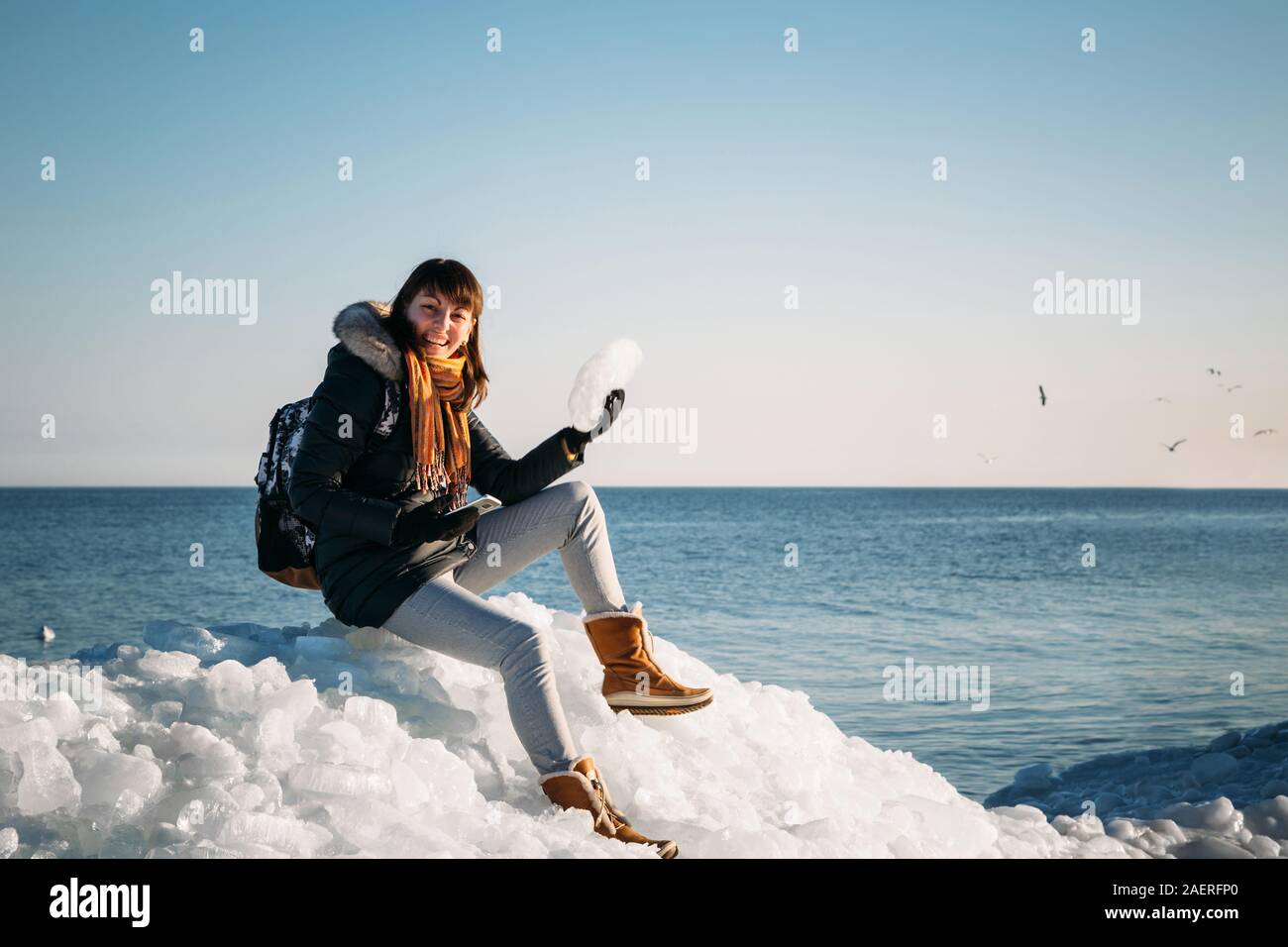 Mujer sonriente joven sentado en la parte superior de los bloques de hielo marino en la costa sujetando la pieza rota de hielo, con un mar azul y el cielo y el fondo. Foto de stock