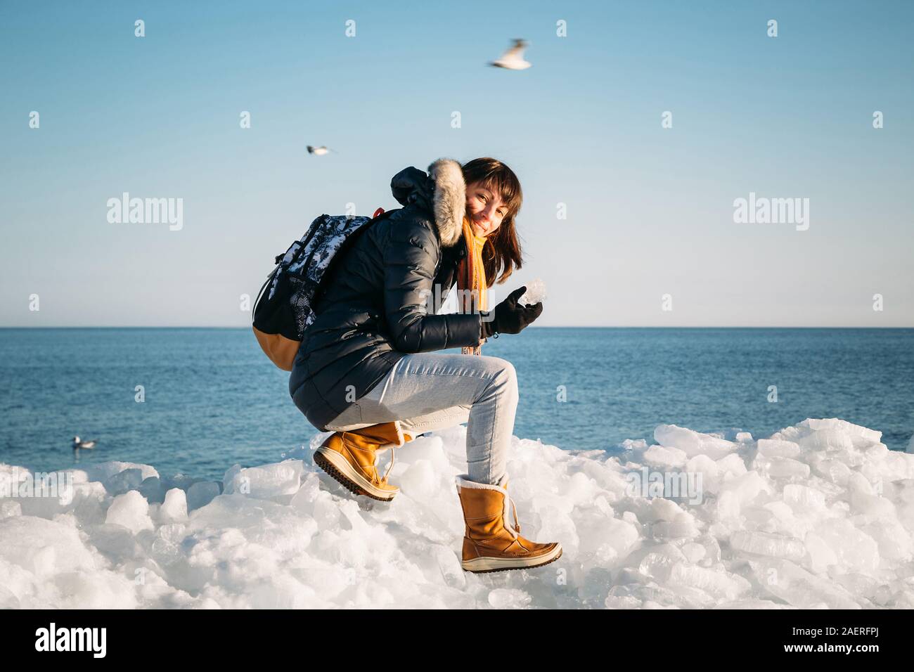 Mujer sonriente joven sentado en la parte superior de los bloques de hielo marino en la costa sujetando la pieza rota de hielo, con un mar azul y el cielo y el fondo. Foto de stock