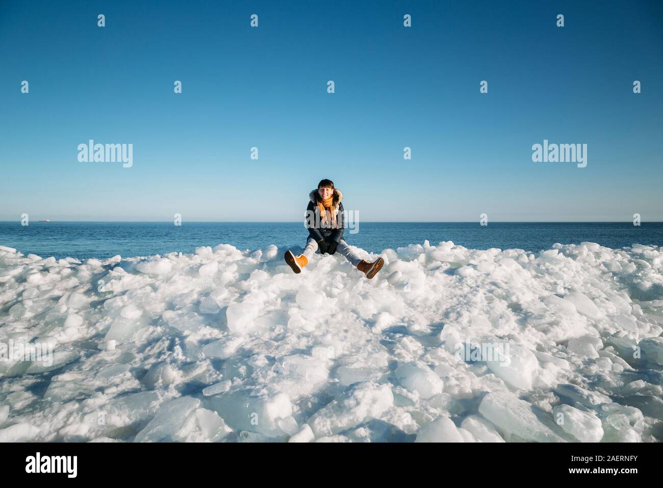 Mujer sonriente joven sentada sobre una cima de mar congelado bloques de hielo en la costa de la mar con un cielo azul en el fondo, en frosty día soleado Foto de stock