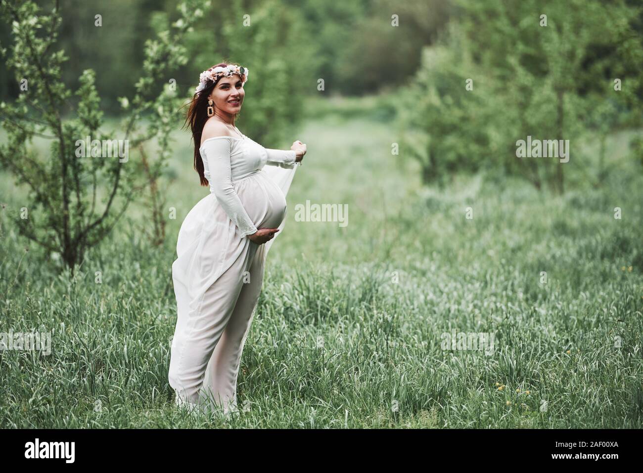 Corona de flores en la cabeza. Hermosa mujer embarazada en vestido tienen una caminata al aire libre. Morena positivo Fotografía de stock -
