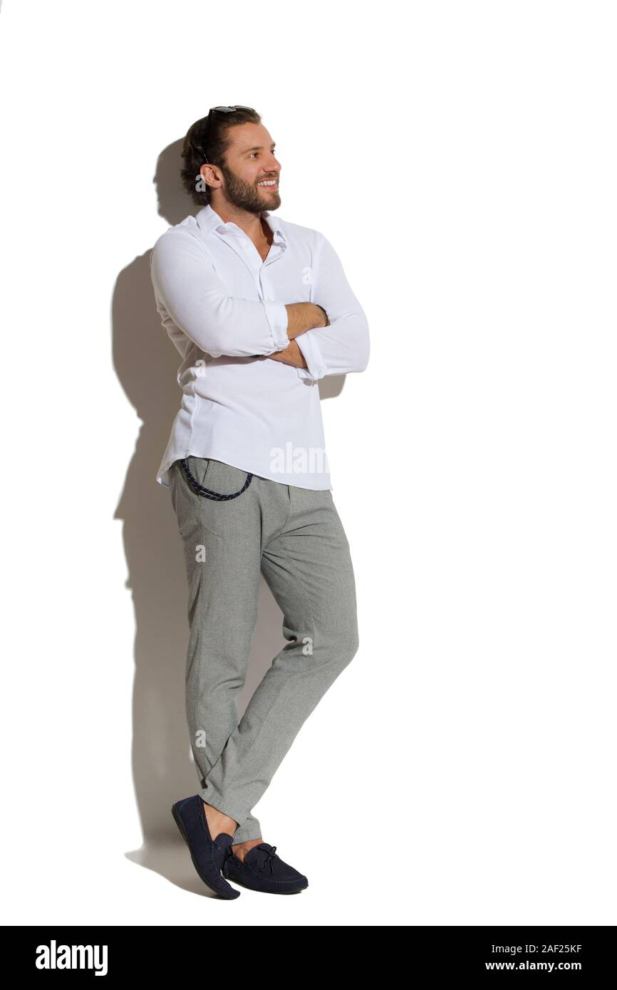 Guapo en los mocasines, pantalón gris y camisa blanca está de pie con los  brazos cruzados, apoyándose en la pared soleada , apartar la mirada y  sonriendo Fotografía de stock - Alamy