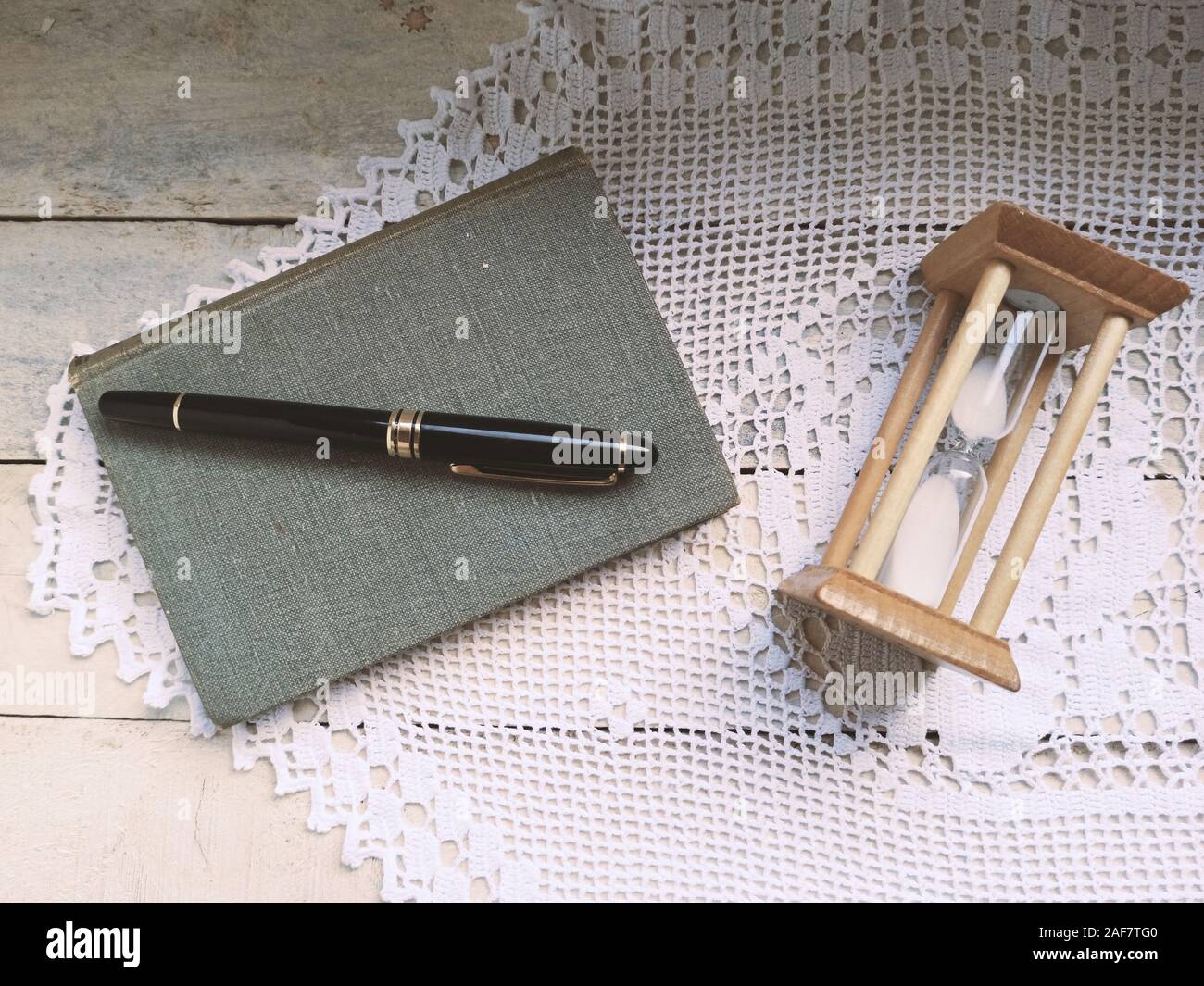 Retro imagen de un reloj de arena en un viejo libro y un lápiz para tomar notas. Mesa de madera, decorado con tapices artesanales crochet. Foto de stock