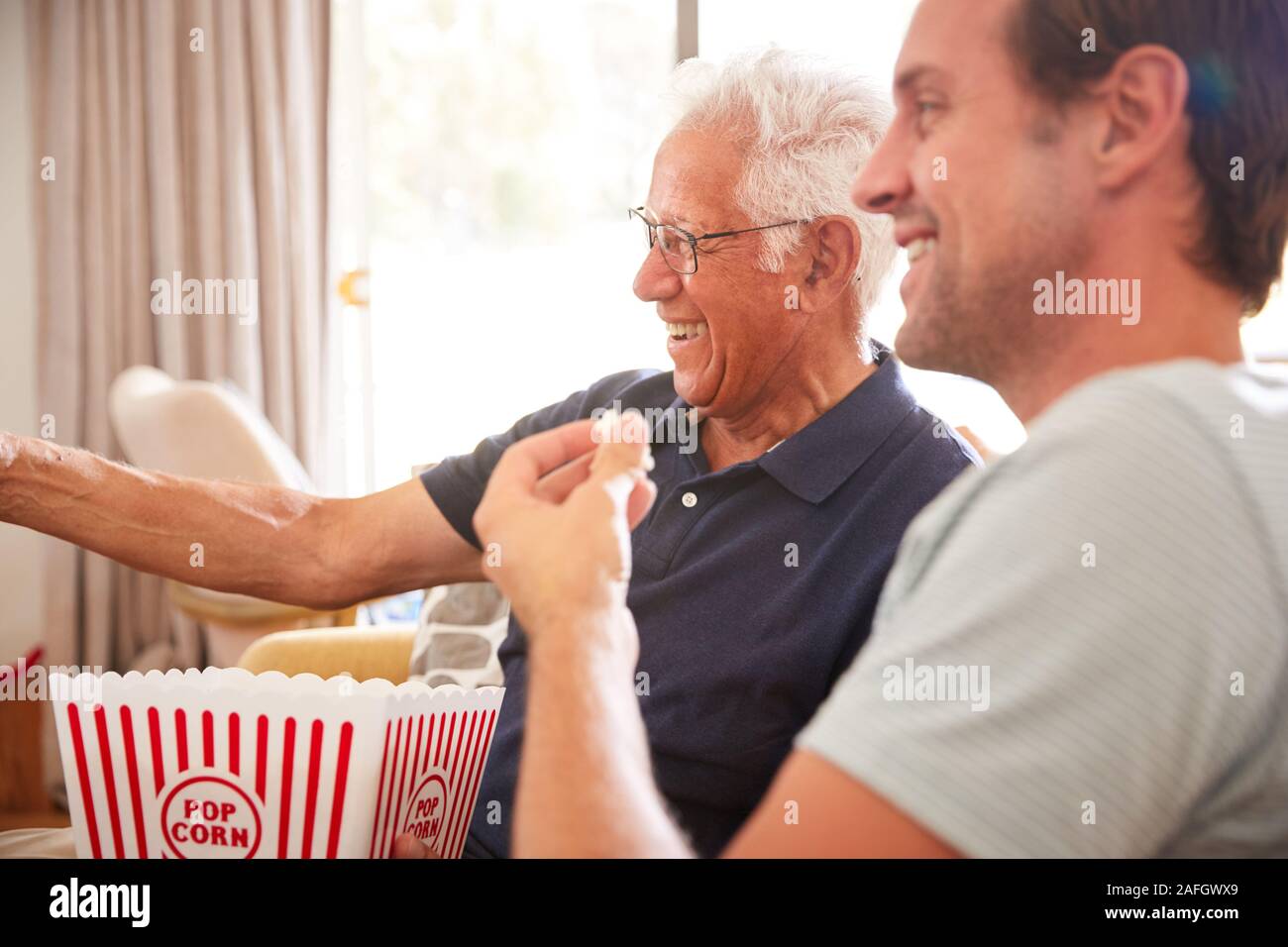 El padre con el hijo adulto comiendo palomitas viendo la película en el sofá en casa juntos Foto de stock