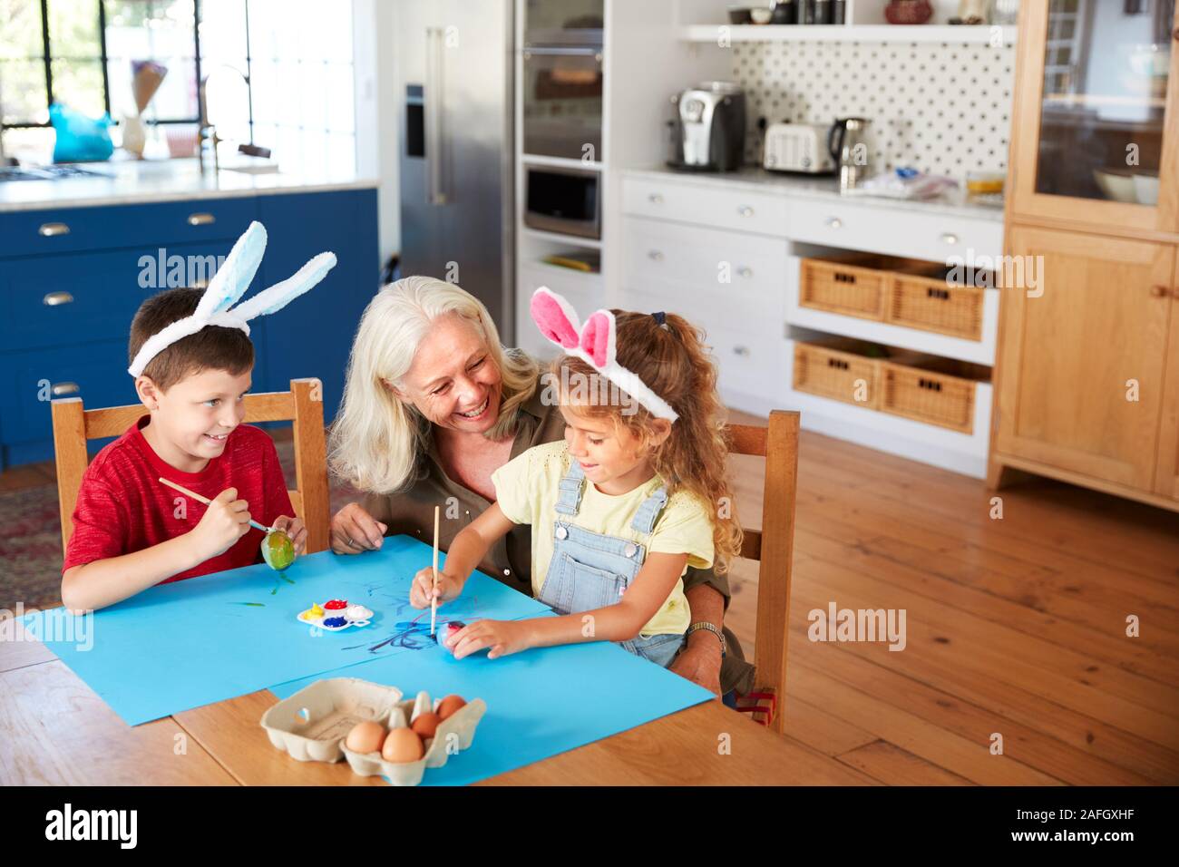 Abuela con nietos vestidos de orejas de conejo decorando huevos de Pascua en el hogar Foto de stock