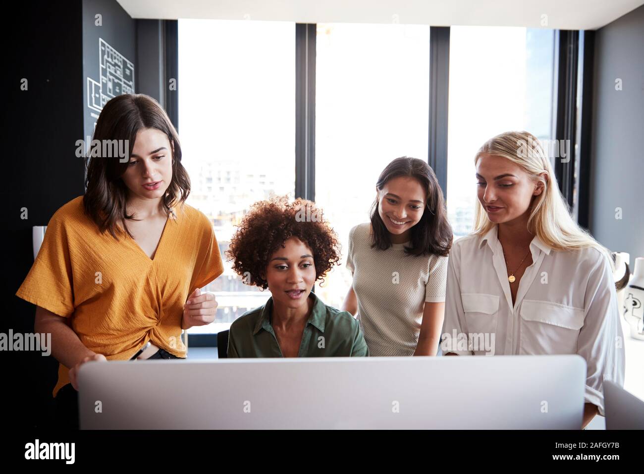 Cuatro mujeres creativos trabajan en torno a un monitor de ordenador en una oficina, vista frontal, cerrar Foto de stock