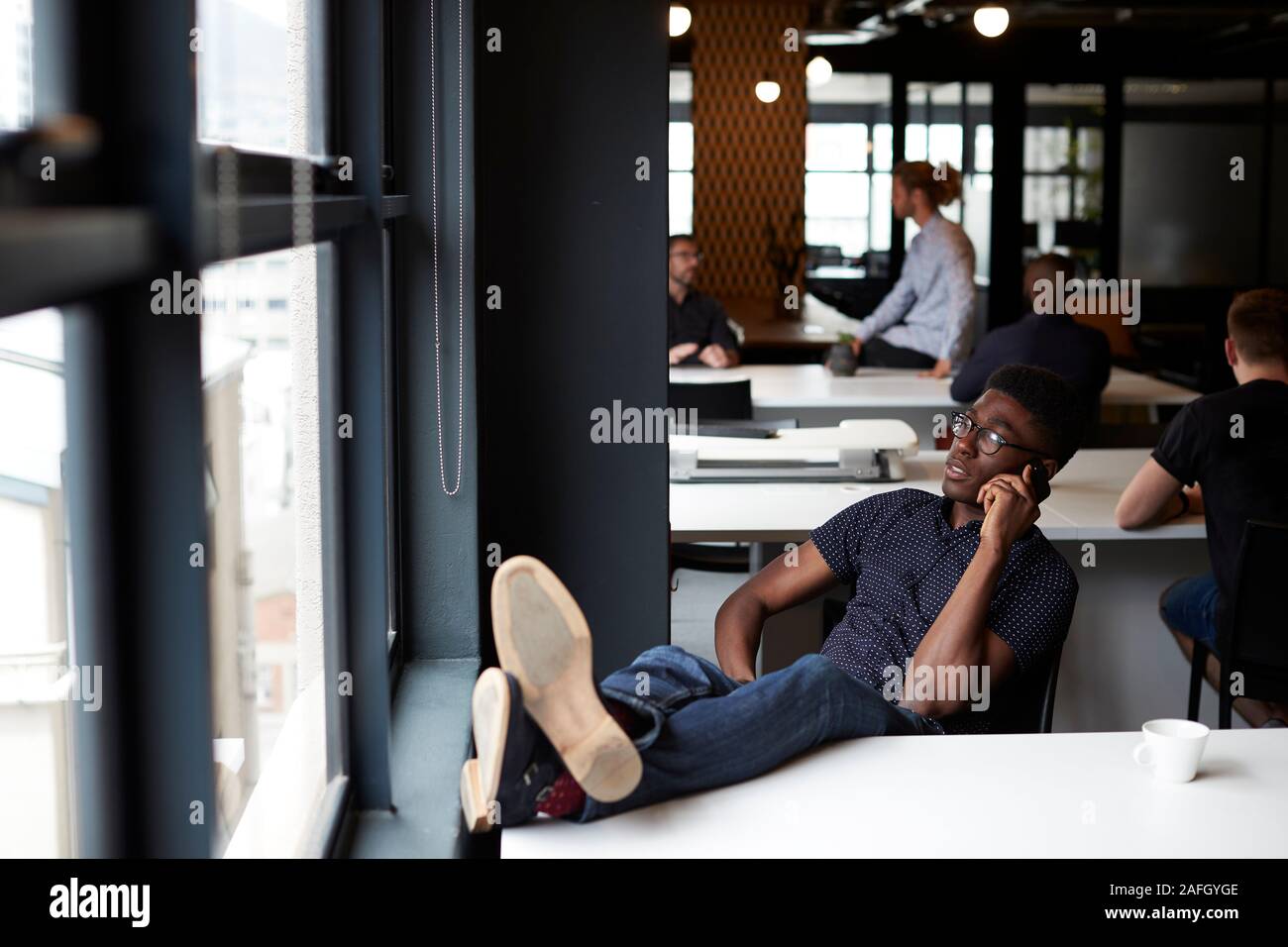 Jóvenes creativos macho negro sentado en una oficina con sus pies sobre el escritorio con teléfono, cerrar Foto de stock