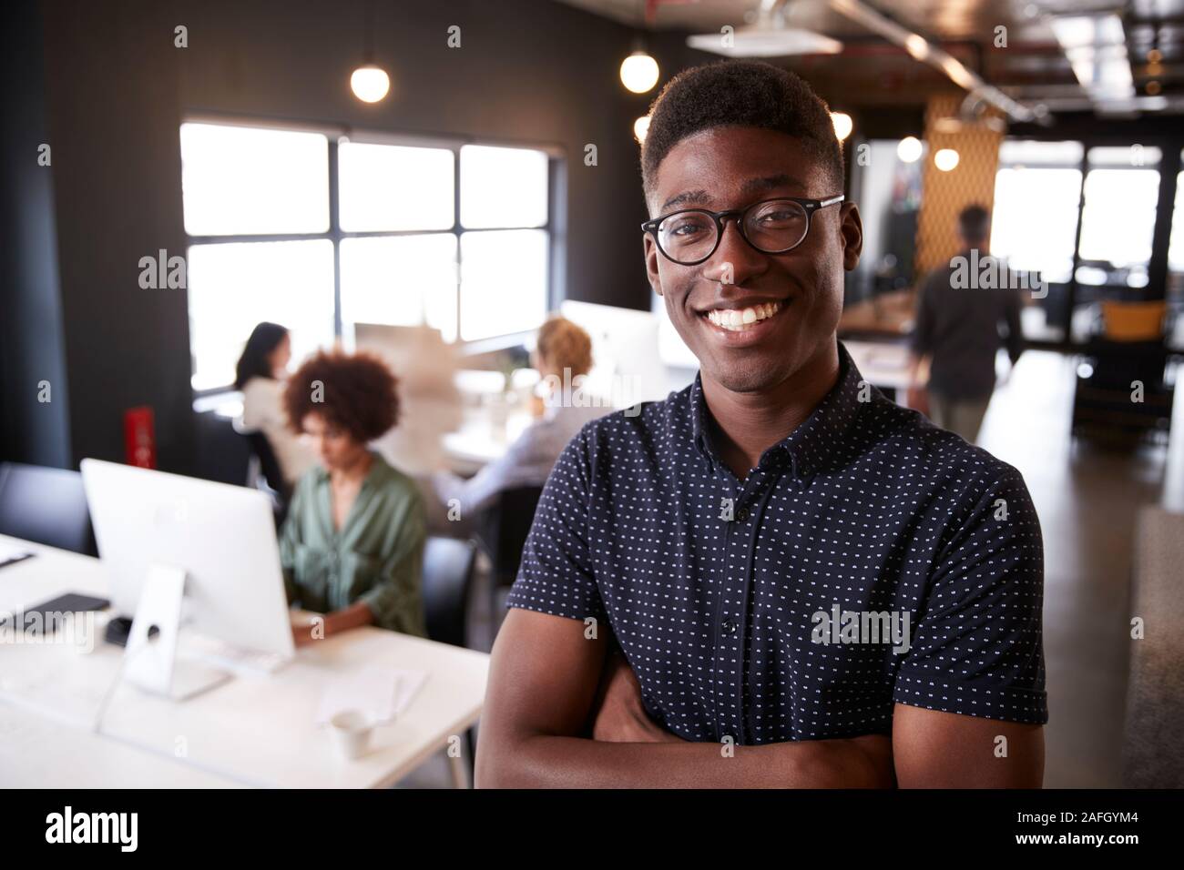 Macho negro milenario permanente creativo en una concurrida oficina informal, sonriendo a la cámara Foto de stock