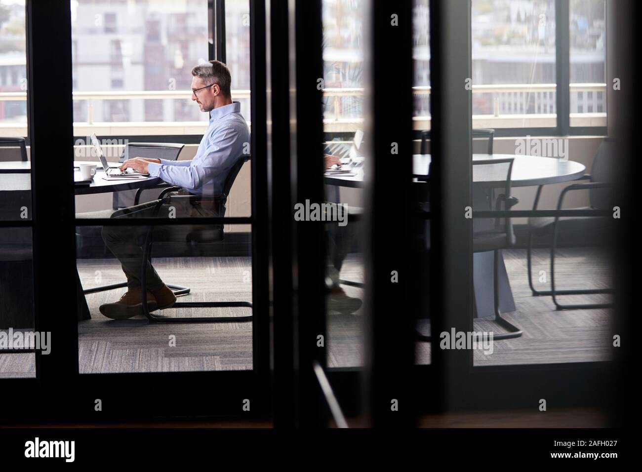 Empresario blanco de mediana edad que trabajan solos en una sala de reuniones, visto a través de la puerta de vidrio Foto de stock