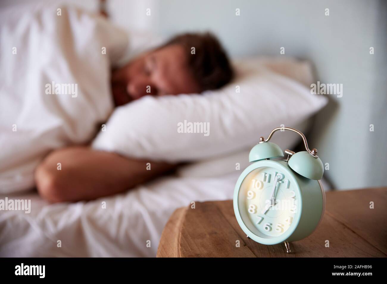 Hombre adulto medio dormido en la cama, despertador en la mesilla de noche en primer plano, se enfocan en primer plano Foto de stock