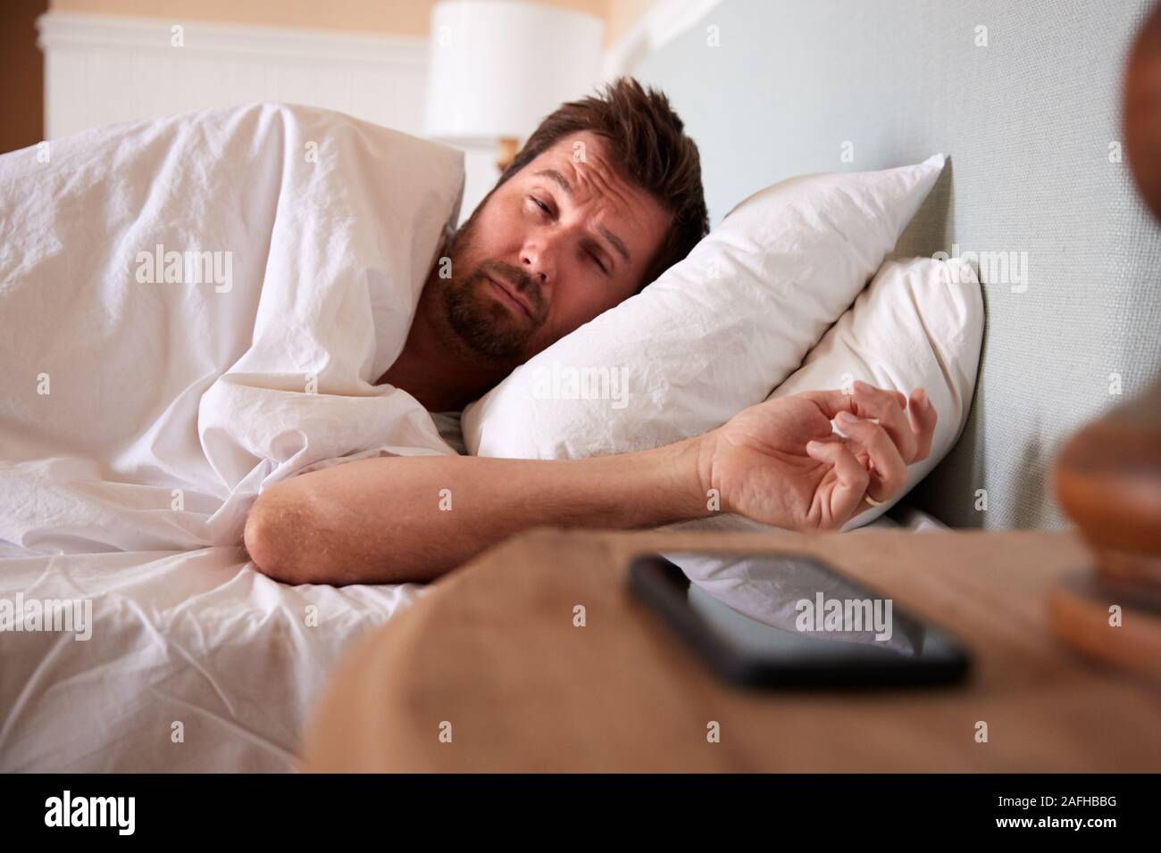 Hombre adulto medio dormido en la cama, mirando el smartphone en la mesilla de noche en primer plano Foto de stock