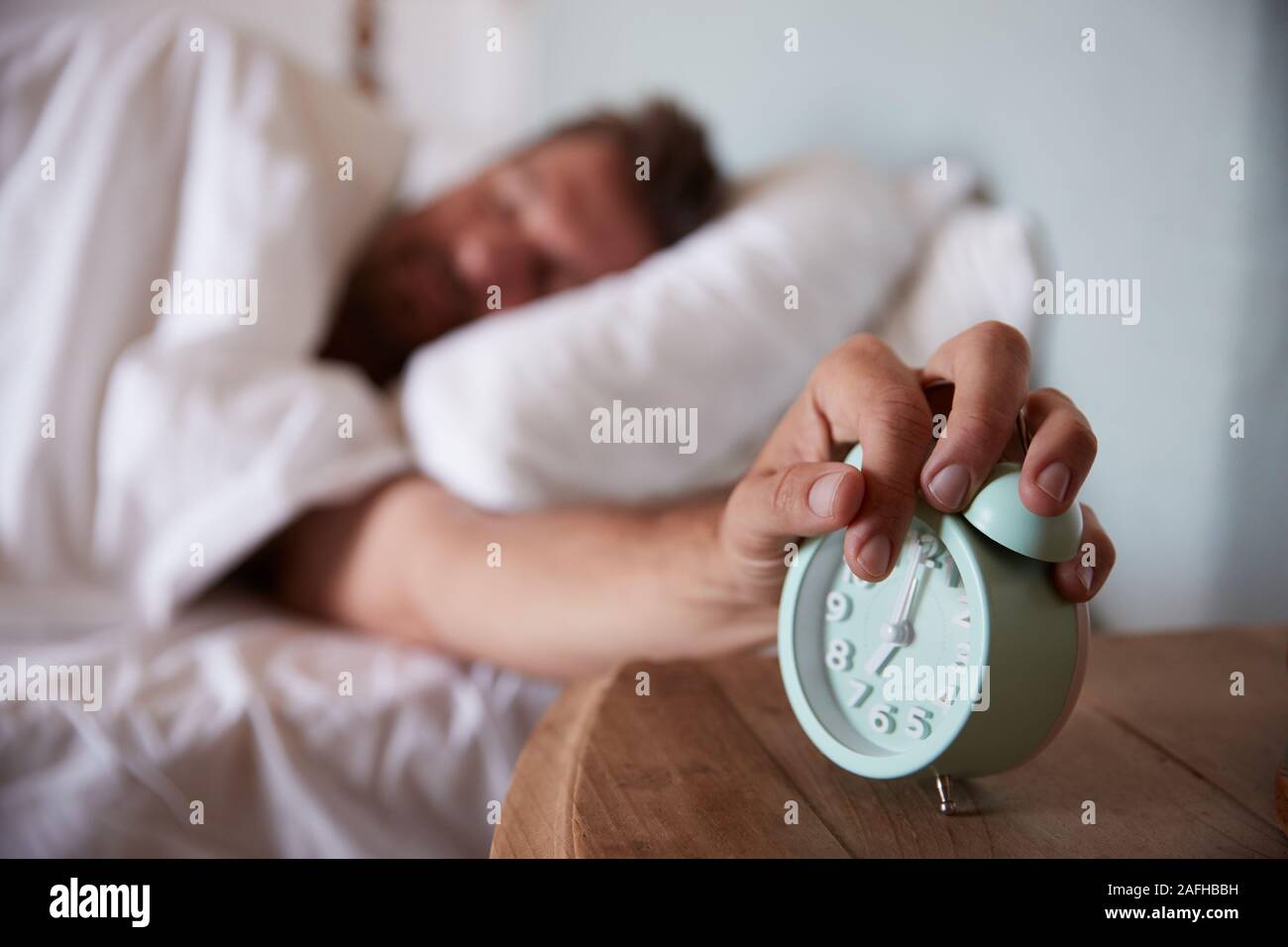 Hombre adulto medio dormido en la cama, llegando a despertador en la mesilla de noche en primer plano Foto de stock