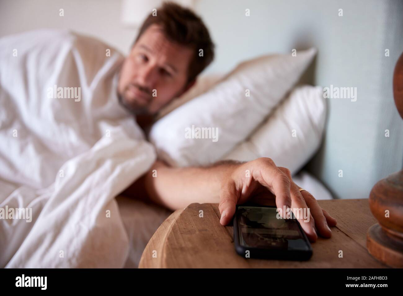 Hombre adulto medio acostado en la cama, llegando al smartphone en la mesilla de noche en primer plano Foto de stock