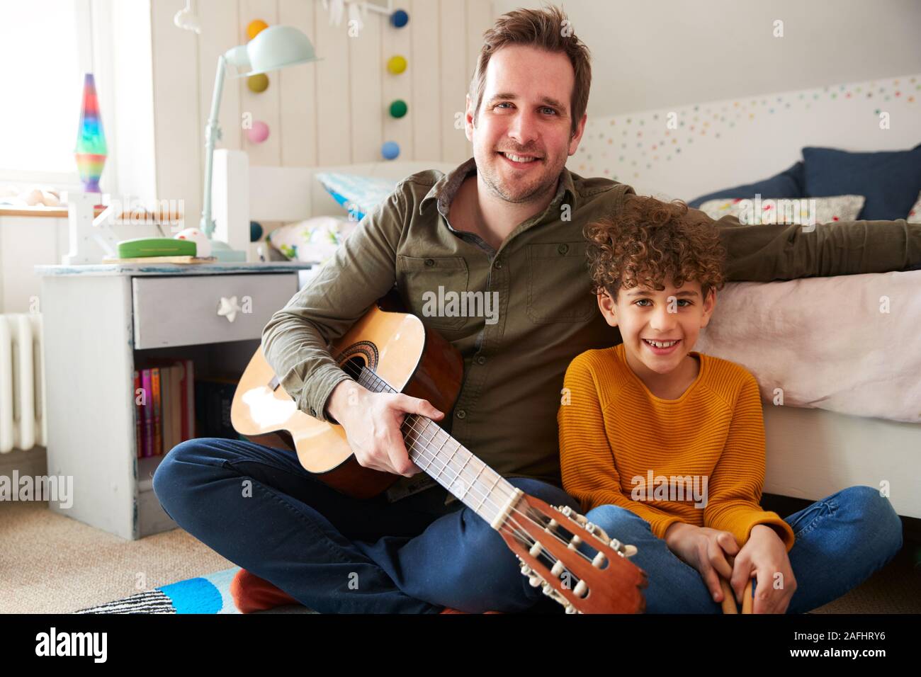 Retrato de un solo padre en casa con su hijo tocando la guitarra acústica en el dormitorio Foto de stock