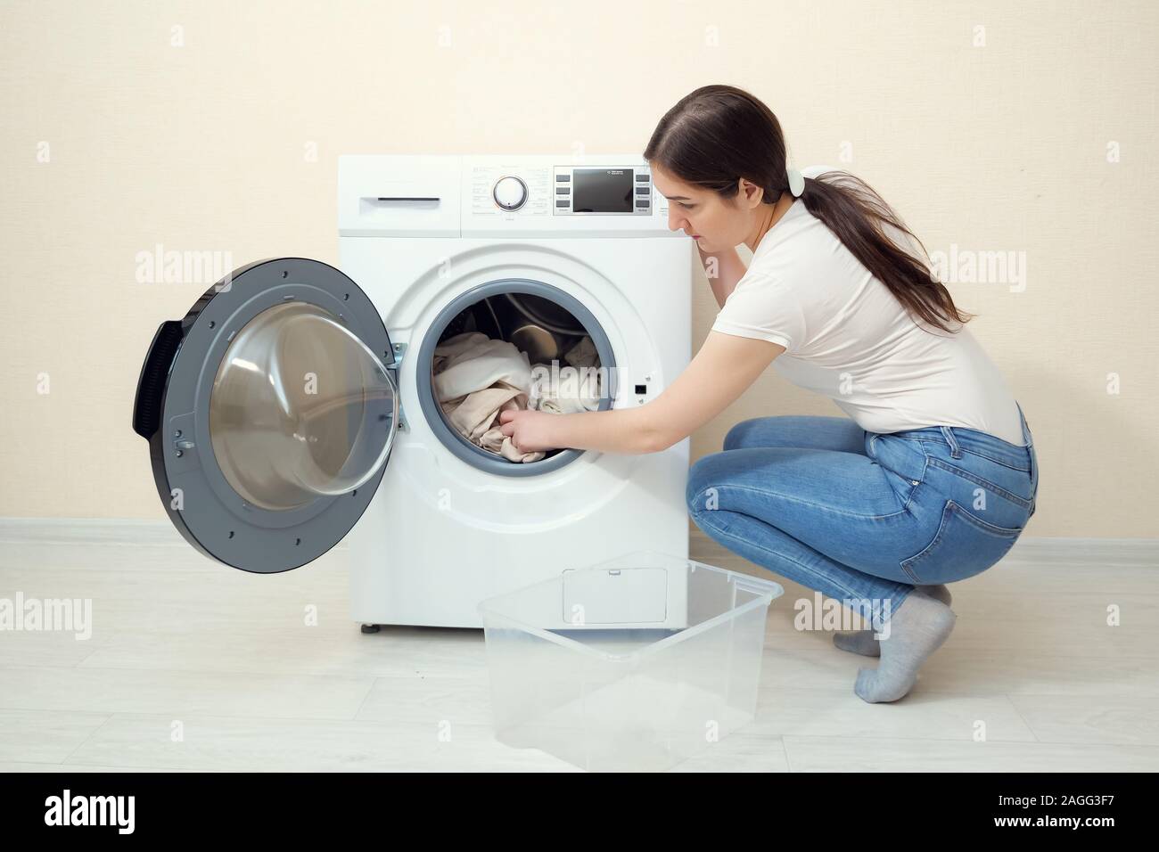 Cargas ama de casa joven con la ropa sucia en la lavadora moderno de caja de plástico cerca de pared beige Fotografía de stock -
