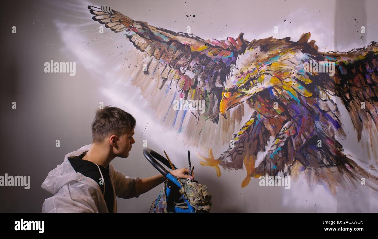 Artista diseñador dibuja un águila en la pared. Artesano decorador pinturas fotografía con acrílico color de aceite. Pintor pintor vestido con capa de pintura. Indoor. Foto de stock
