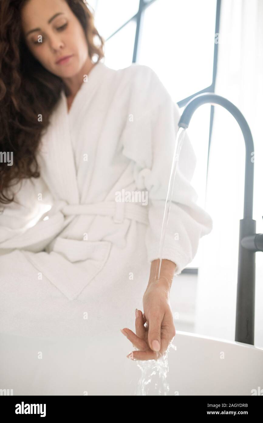 Chica en albornoz blanco manteniendo su mano bajo corriente de agua que fluye en el grifo. Foto de stock