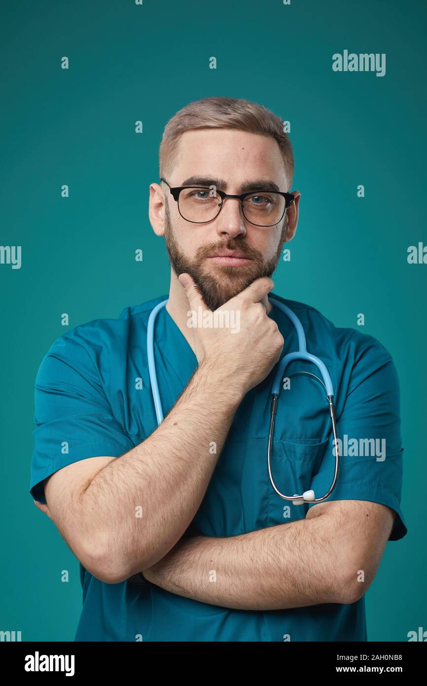 Cintura para arriba vertical studio retrato de pensive adulto joven doctor vistiendo uniforme azul y anteojos tocando su barbilla pensando en algo Foto de stock