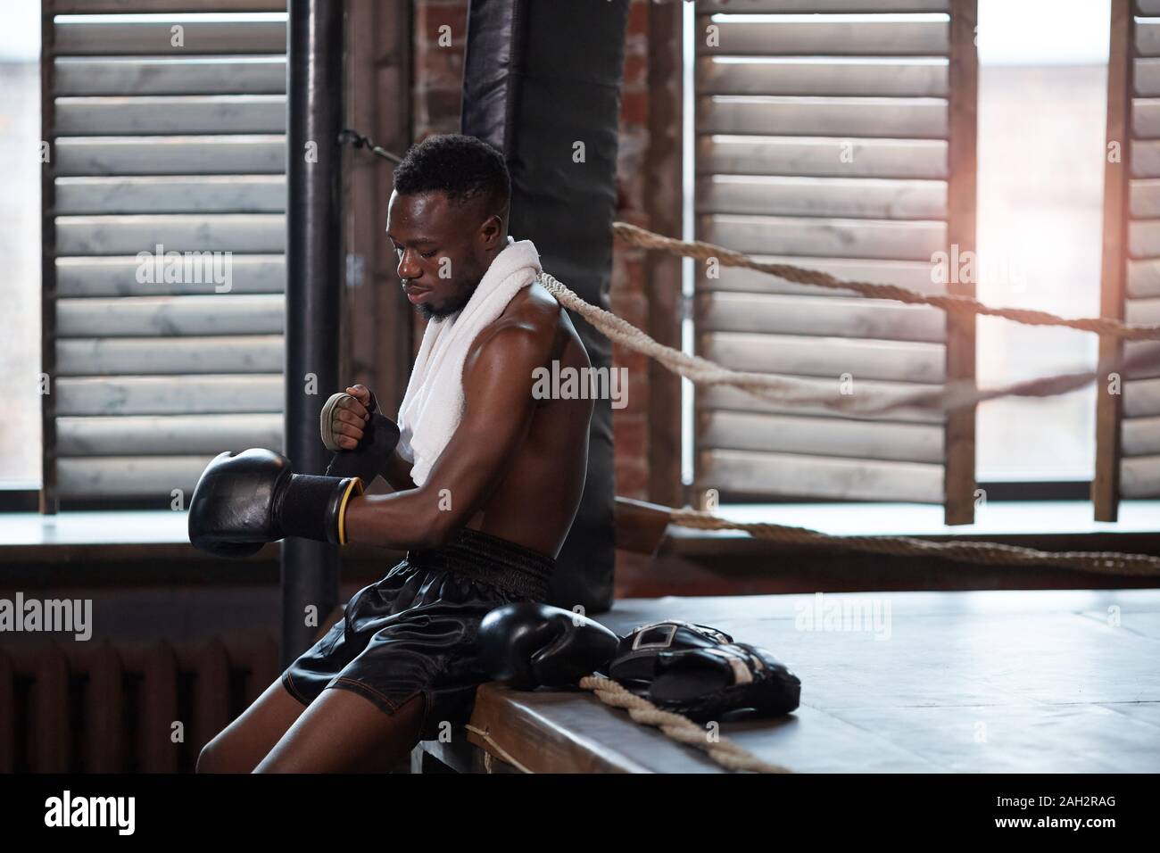 Boxer muscular africana sentada cerca del cuadrilátero vistiendo guantes de boxeo se prepara para luchar Foto de stock