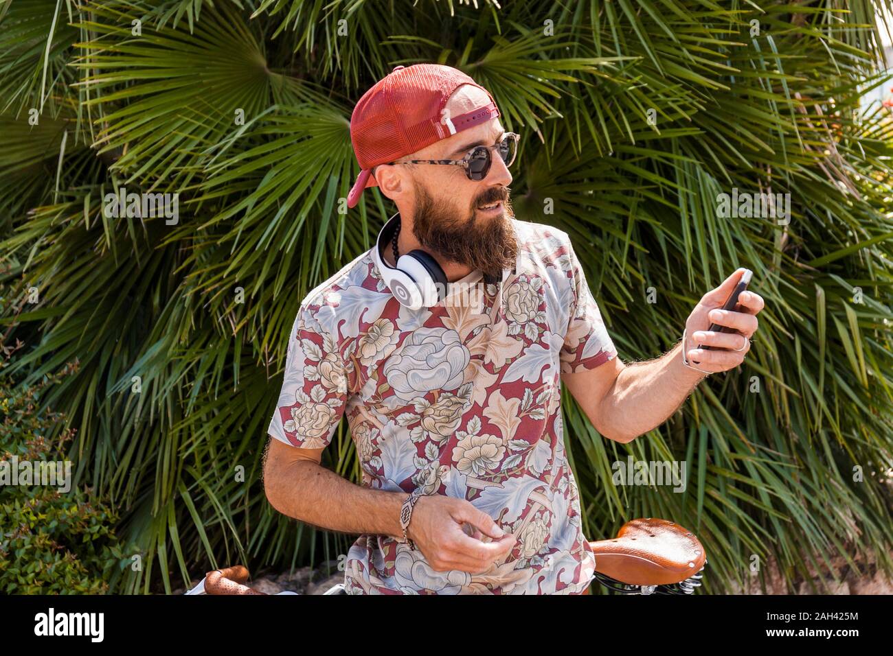 Hombre maduro con rojo basecap, gafas de sol y auriculares blancos usando el smartphone Foto de stock