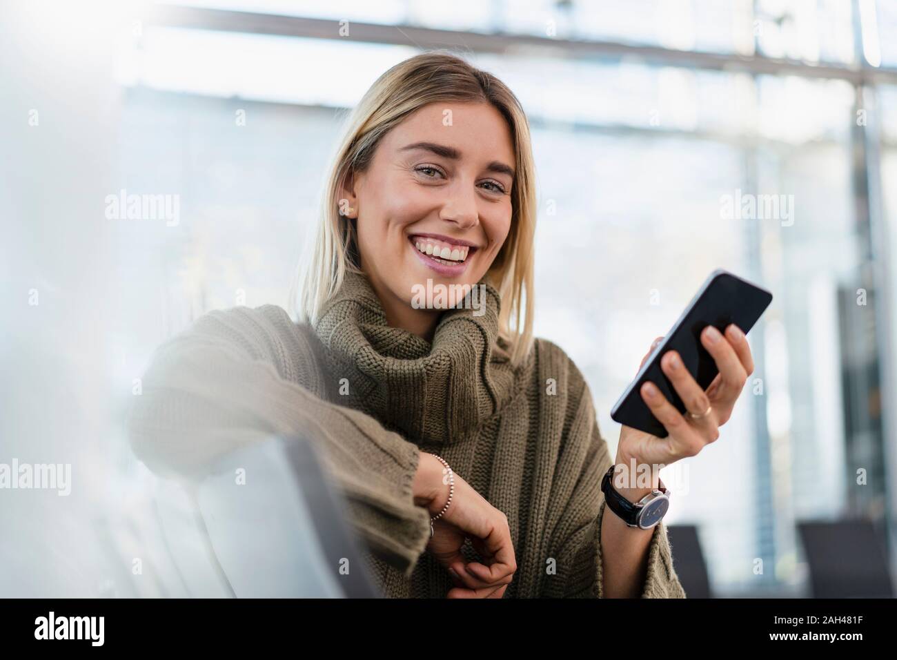 Retrato de mujer joven feliz sentado en el área de espera con teléfono celular Foto de stock