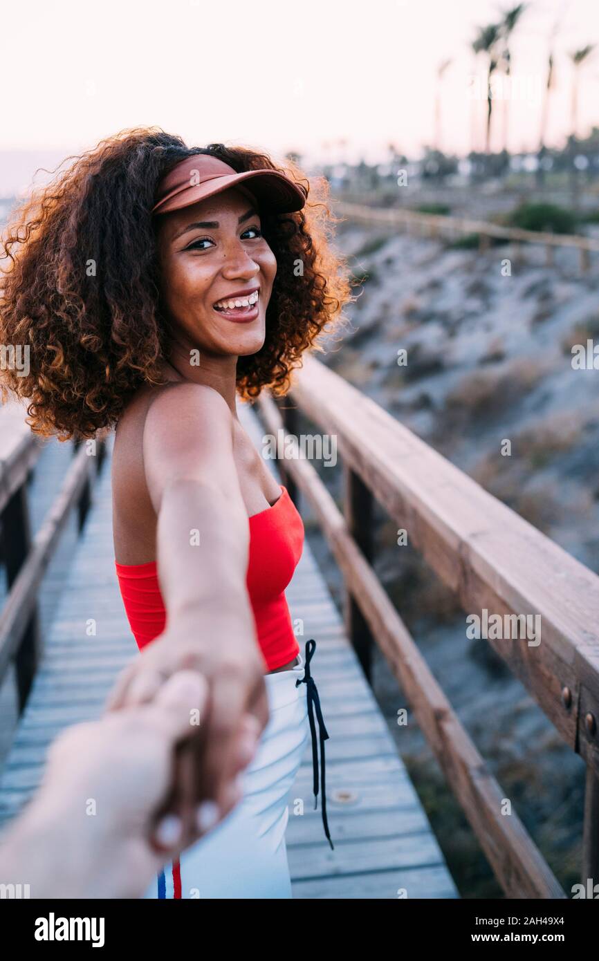 Sonriente joven mujer vistiendo tapa marrón está dirigiendo su mejor amigo a la playa Foto de stock
