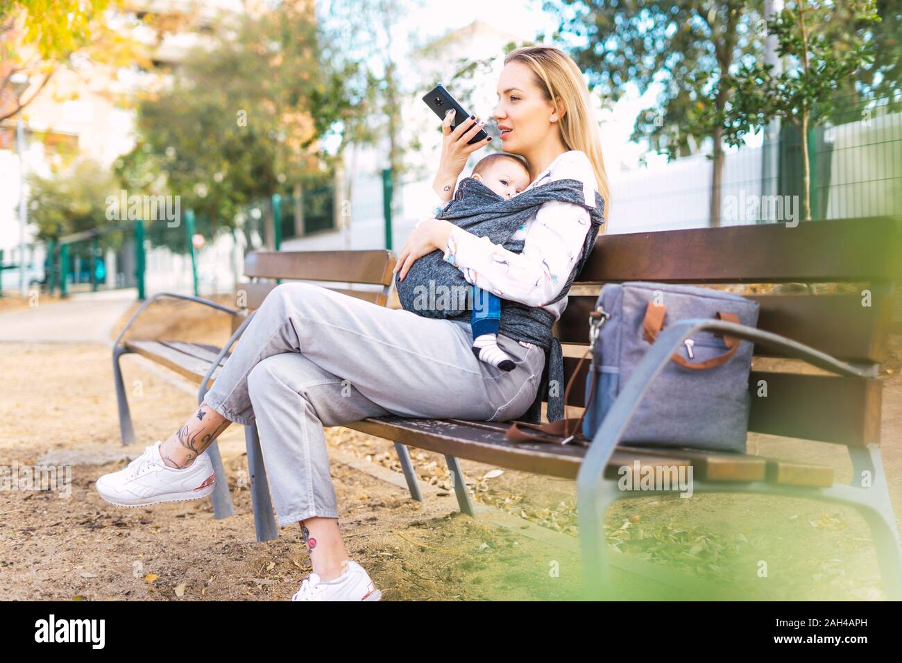 Madre descansando con Baby Boy en una banca del parque utilizando el smartphone Foto de stock