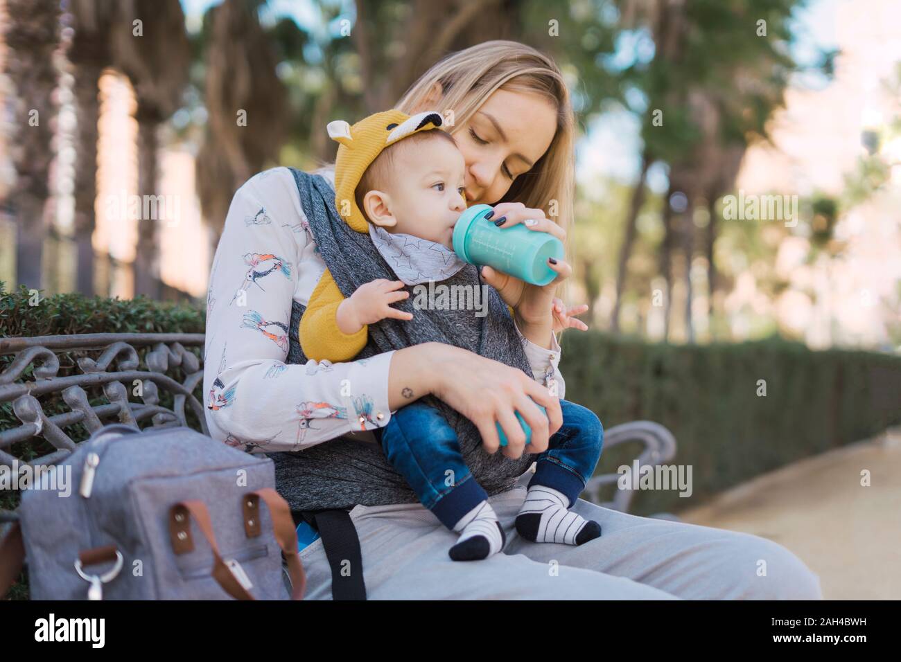 Madre biberón Baby Boy en una banca del parque Foto de stock