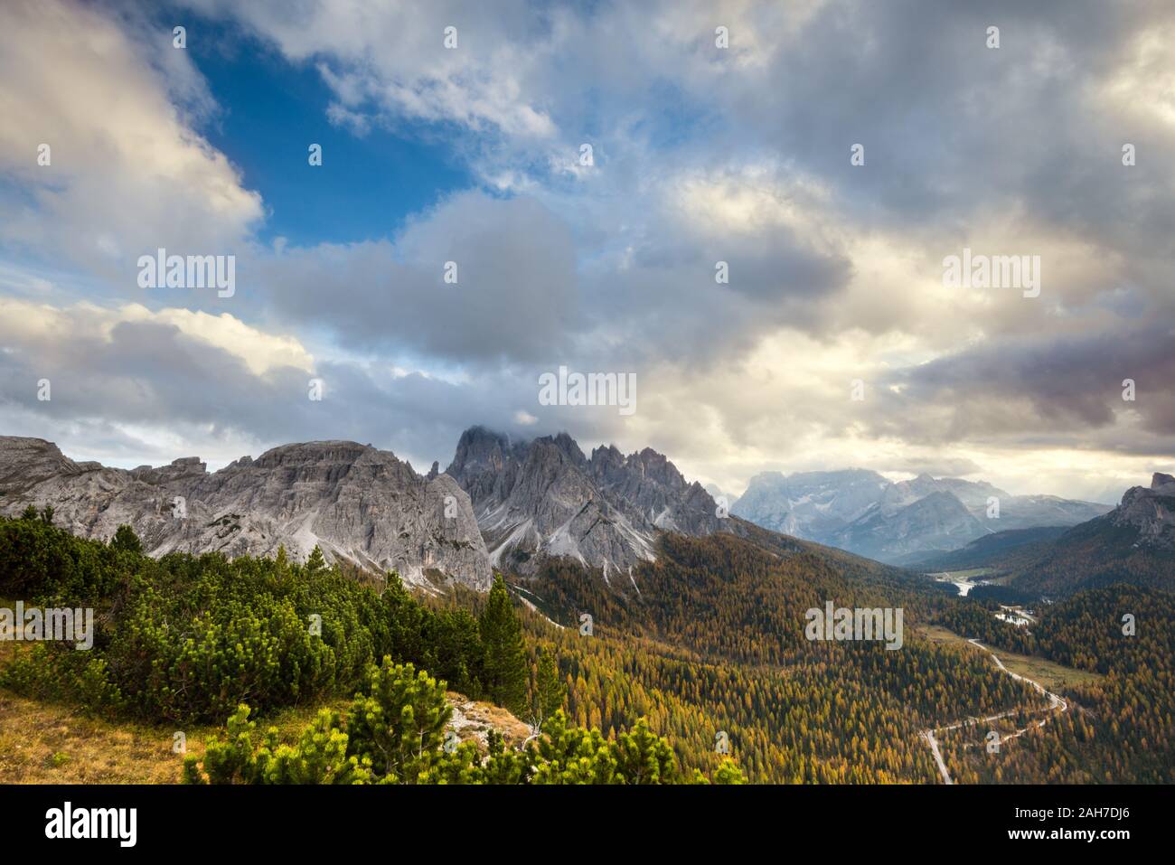 Vista angular de un icónico paisaje otoñal italiano, con valles cubiertos de pinos y montañas distantes, bajo un cielo azul con nubes hinchadas Foto de stock