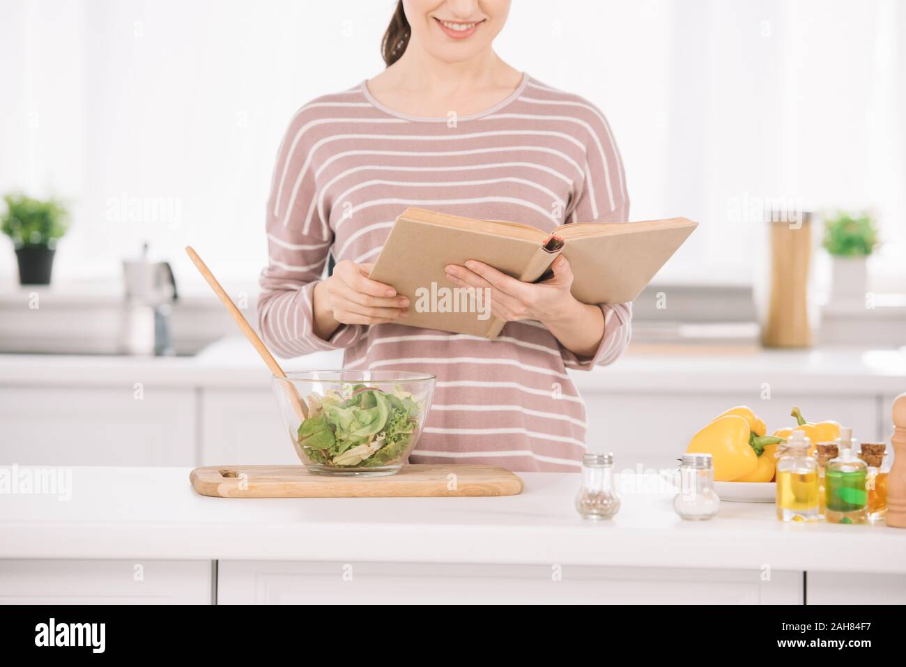 Vista recortada de mujer sonriente lectura libro recibo mientras estuve en la mesa de la cocina cerca del recipiente con ensalada de verduras Foto de stock