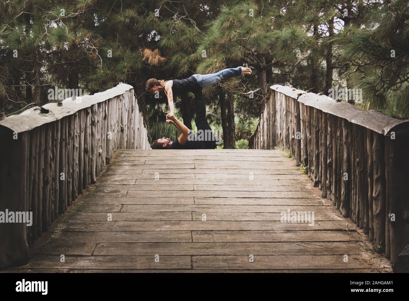 Hombre y mujer en ropa urbana practicando yoga acro en puente de madera en el parque. Yogui par en equilibrio plantean al aire libre. Efecto de tono oscuro Foto de stock