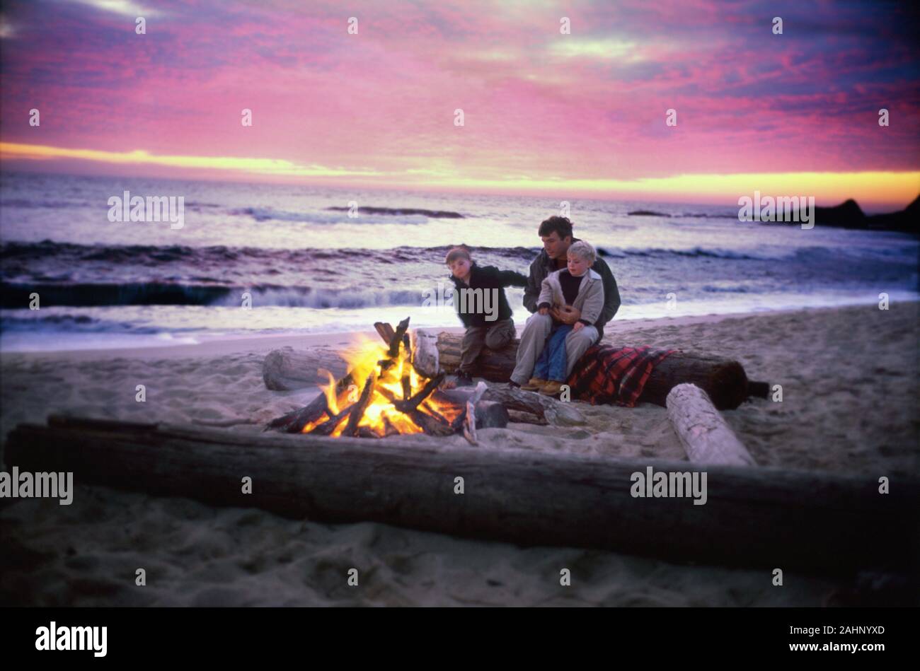Un padre se sienta frente a un fuego con sus hijos en una playa pintoresca. Foto de stock