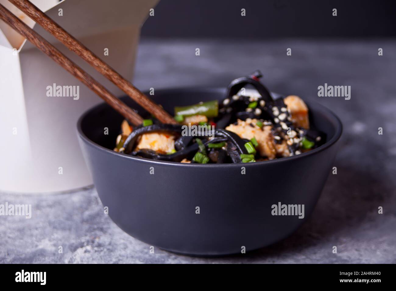 Tinta de calamar negro fideos udon con carne de pollo en el recipiente negro con palillos sobre fondo de piedra oscura y caja de papel cerca. Saque el concepto de alimentos Foto de stock