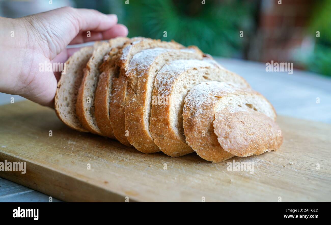 Mano sujetando una hogaza de rodajas de pan de masa fermentada en placa de madera Foto de stock