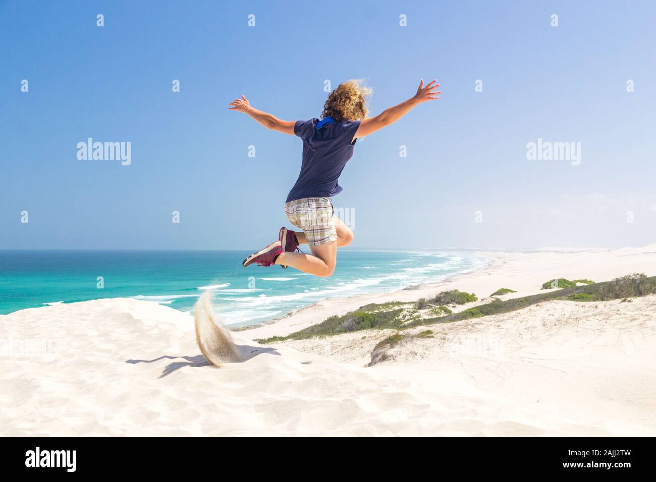 Mujer joven saltando de una duna de arena, vista al mar turquesa y las blancas arenas de la costa, la reserva natural De Hoop, Sudáfrica Foto de stock