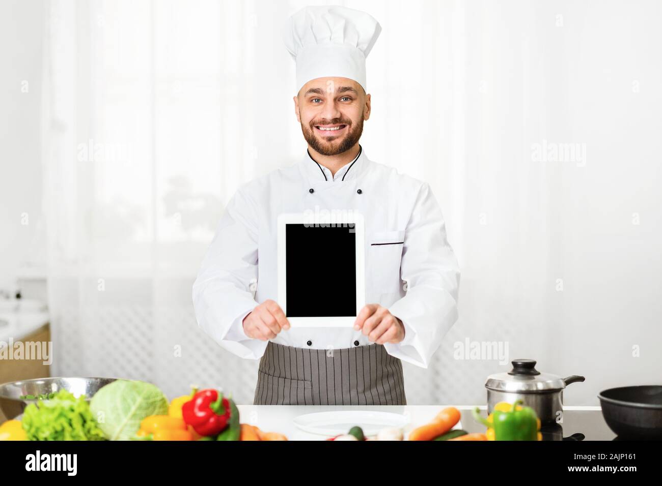 Cocinar App. Chef hombre mostrando tableta digital con pantalla vacía o Restaurante Menú publicidad permanente de aplicación en interior de la cocina. Boceto Foto de stock
