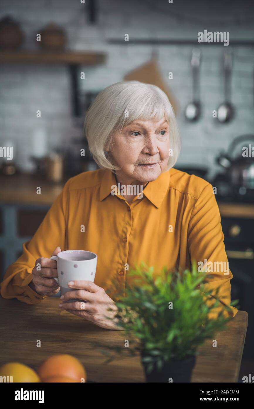Buena anciana en amarillo sentada en su cocina Foto de stock