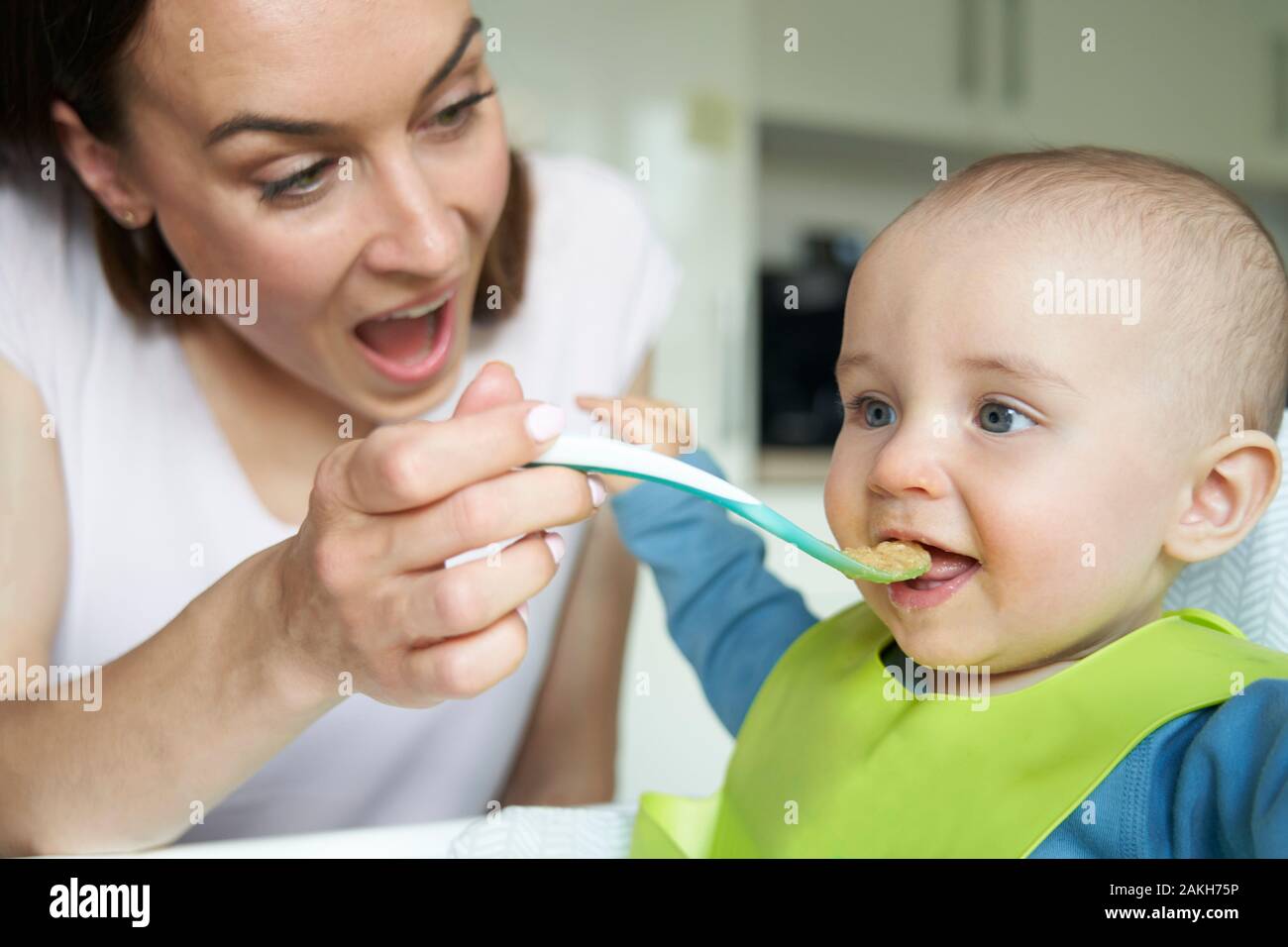 Bebé de 8 meses sonriente niño en casa en alta silla alimentados con alimento sólido por la madre con una cuchara Foto de stock