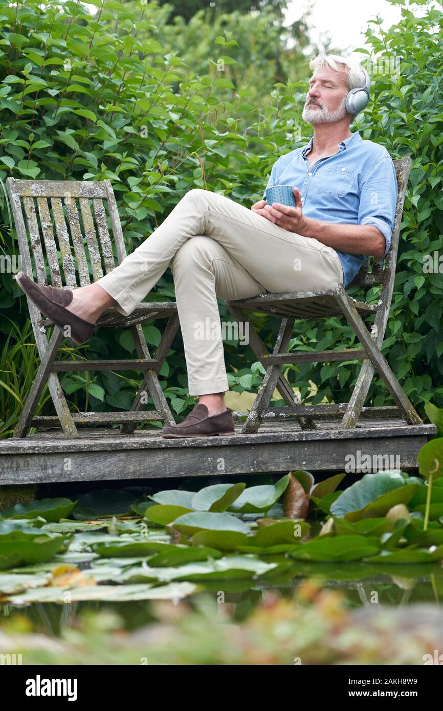 Hombre maduro, relajarse en el jardín escuchando música en auriculares inalámbricos con embarcadero en el lago Foto de stock