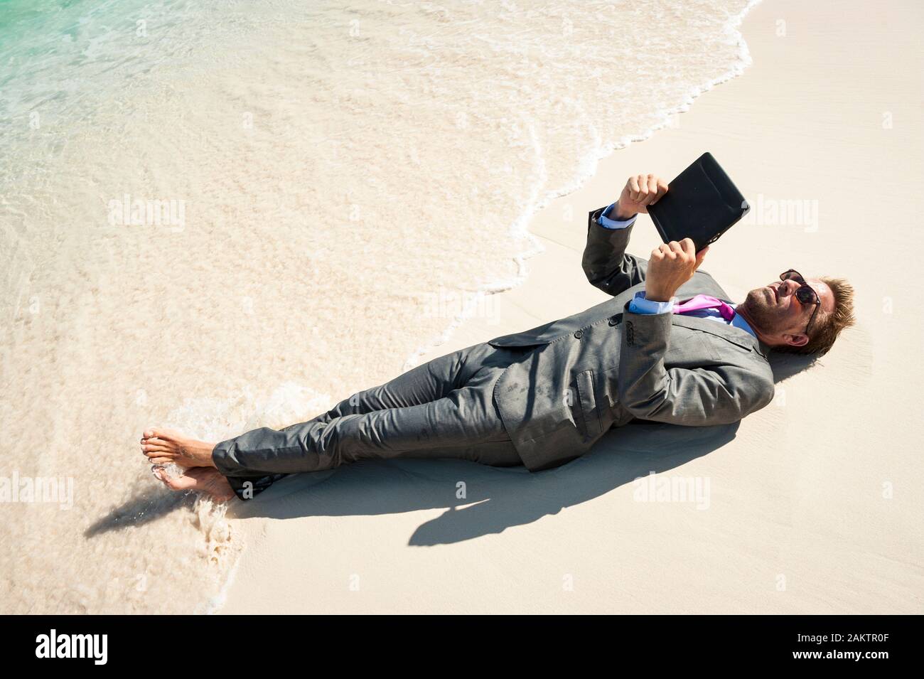 Hombre de negocios relajado acostado en un traje húmedo utilizando su tablet ordenador en la orilla de una playa tropical Foto de stock