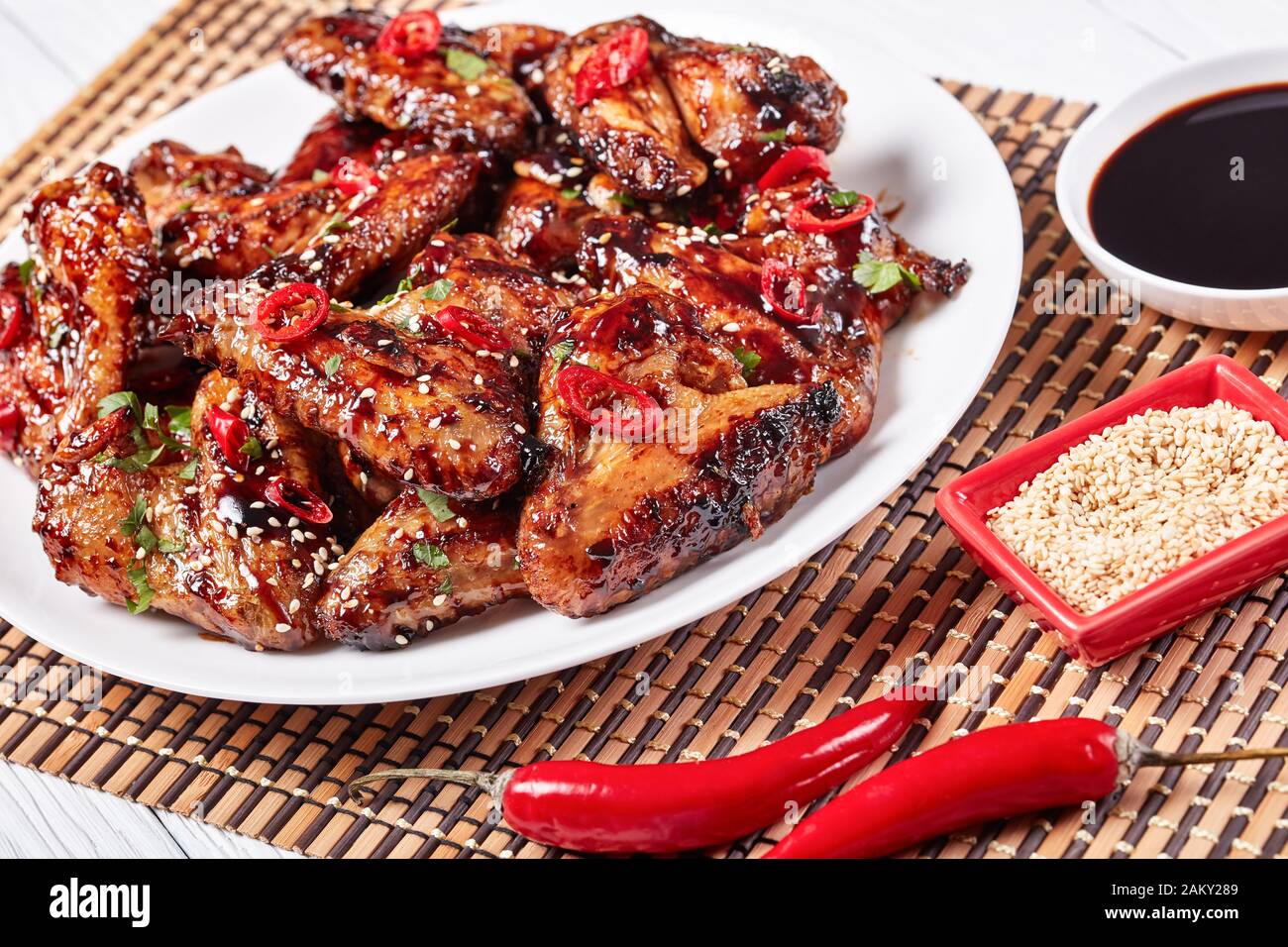 Primer plano de las crujientes alitas de pollo Teriyaki rociadas con  semillas de sésamo, trozos de Chile y perejil en un plato blanco sobre una  alfombra de bambú, vista desde ab Fotografía