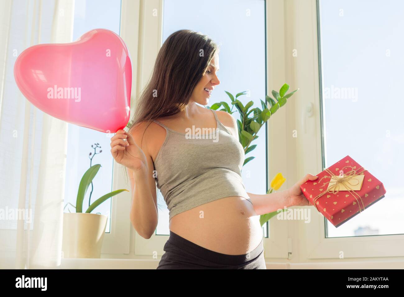 Feliz joven hermosa mujer embarazada con caja de regalo y globo de corazón  rojo, regalo para el día de San Valentín, para las vacaciones de primavera.  Maternidad y embarazo Fotografía de stock -
