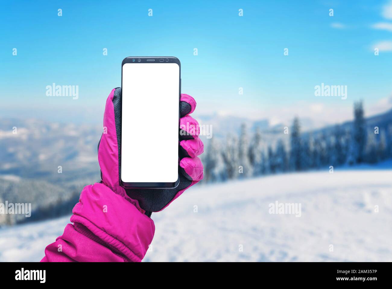 Smartphone se burla en la mano chica con guante. Concepto de usar un teléfono en una estación de esquí. Invierno, con nieve de montaña en el fondo. Guante rosa y j Foto de stock