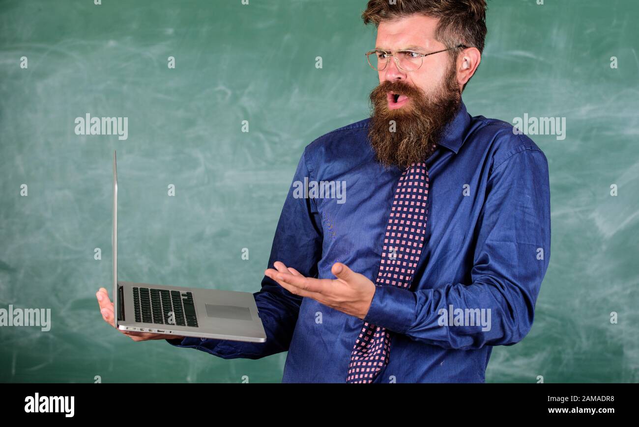 El hombre barbudo del profesor trabajo confuso con el fondo moderno de la pizarra del portátil. Hipster maestro de expresión confusa tiene portátil. Temas de educación a distancia. Temas de enseñanza utilizando tecnologías modernas. Foto de stock